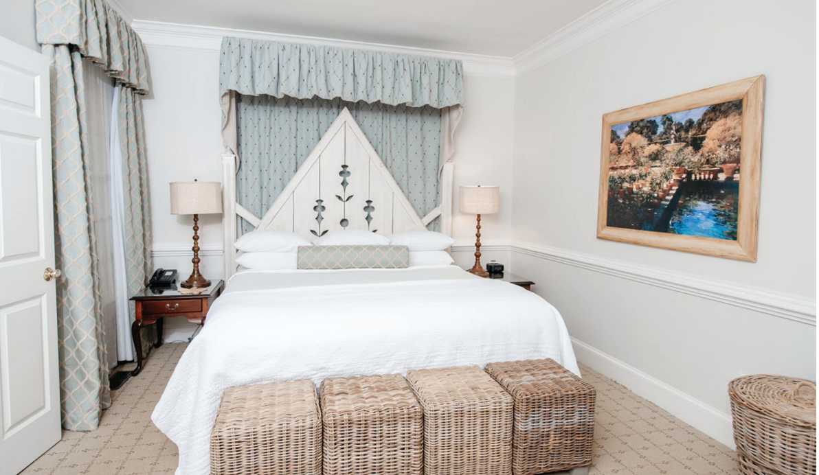 A cozy bedroom with a white bed, flanked by two wooden nightstands with lamps. Three woven wicker storage baskets are at the foot of the bed. A framed landscape painting hangs on the right wall, and light blue drapes adorn the headboard and windows. Fearrington Village